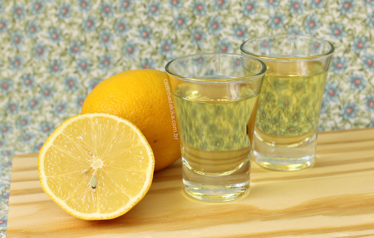 Dois copos de vidro com licor, ao lado de um limão cortado ao meio, sobre uma tábua de madeira.