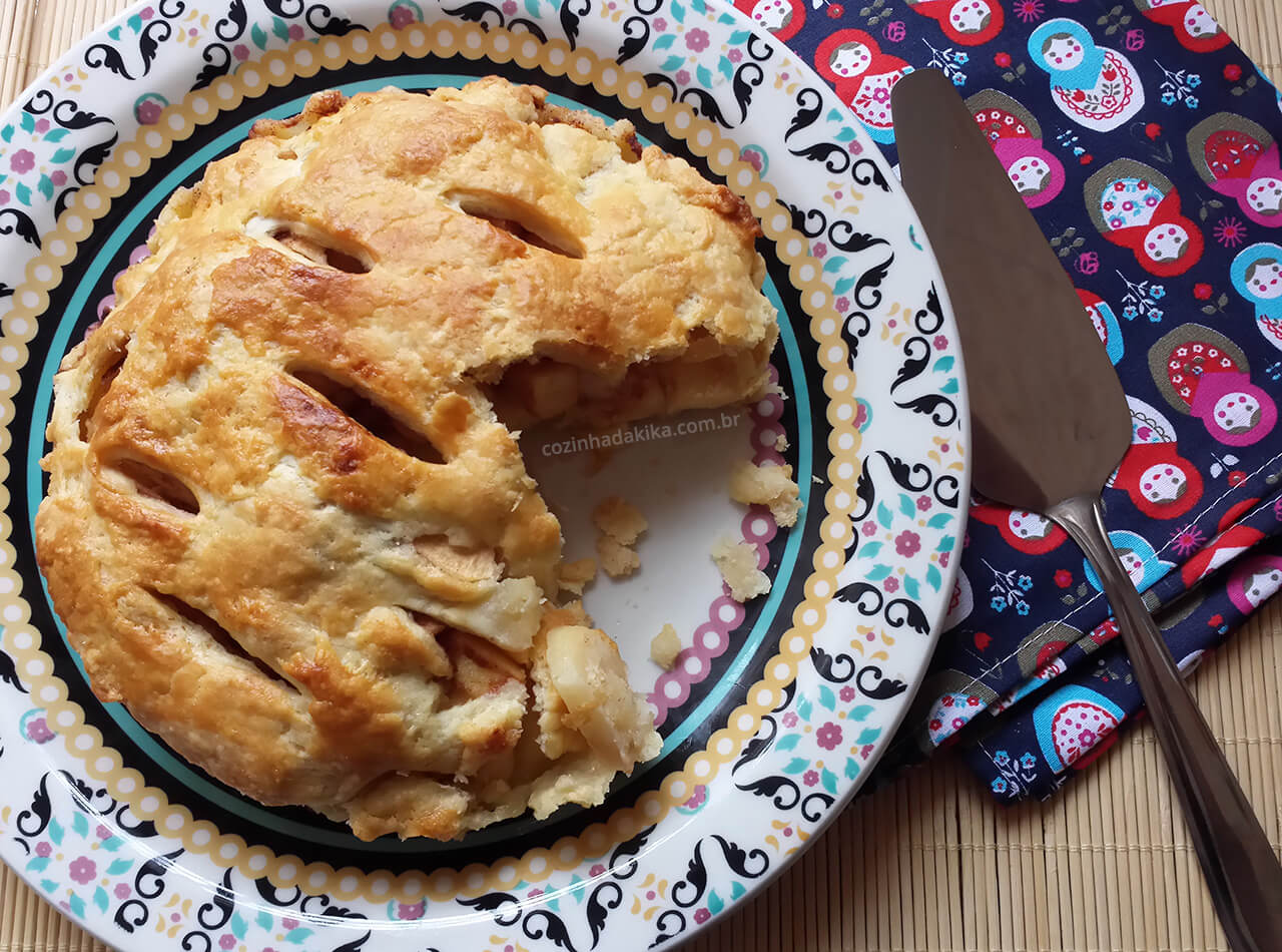 Torta de maçã vista de cima, faltando uma fatia. A torta está num prato estampado, sobre um tecido com estampas de matrioskas. Ao lado do prato, uma espátula.