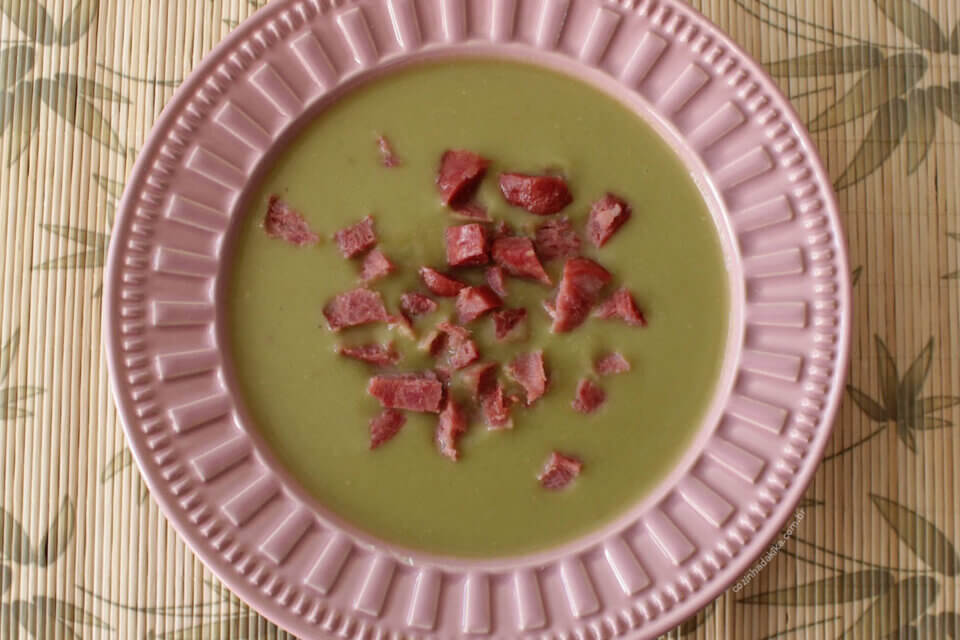 Sopa de ervilha com pedaços de calabresa. O prato é rosa, tem a borda com uma textura decorativa e está sobre uma tábua de bambu.