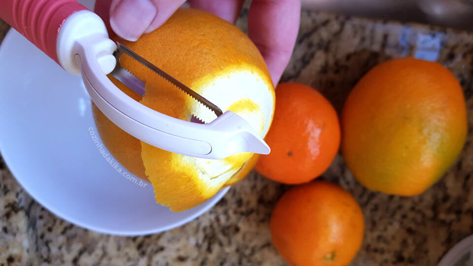 Laranjas sobre uma pia. Uma das laranjas está sendo descascada com um descascador que tira apenas uma camada fina.