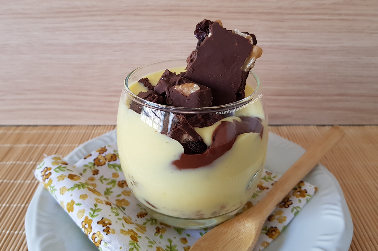 Copo de vidro com um creme de baunilha e pedaços de chocolate com castanhas. O copo está dentro de um prato branco, com uma colher de madeira ao lado, sobre um tecido florido.