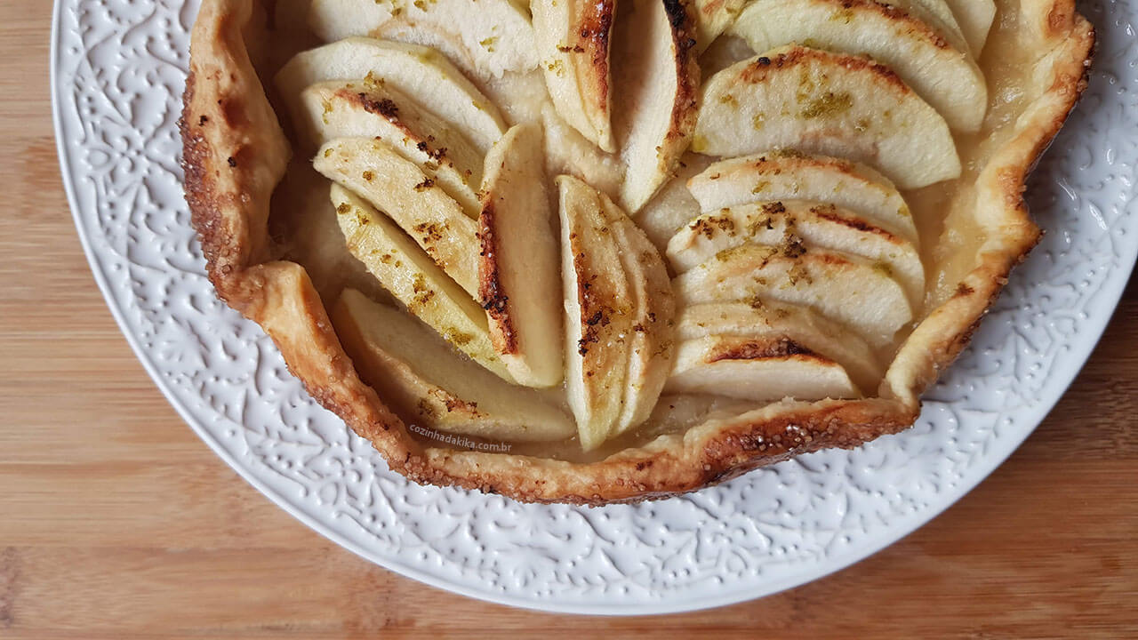 Torta rústica de maçã vista de cima, dentro de um prato branco com a borda trabalhada, sobre uma tábua de madeira